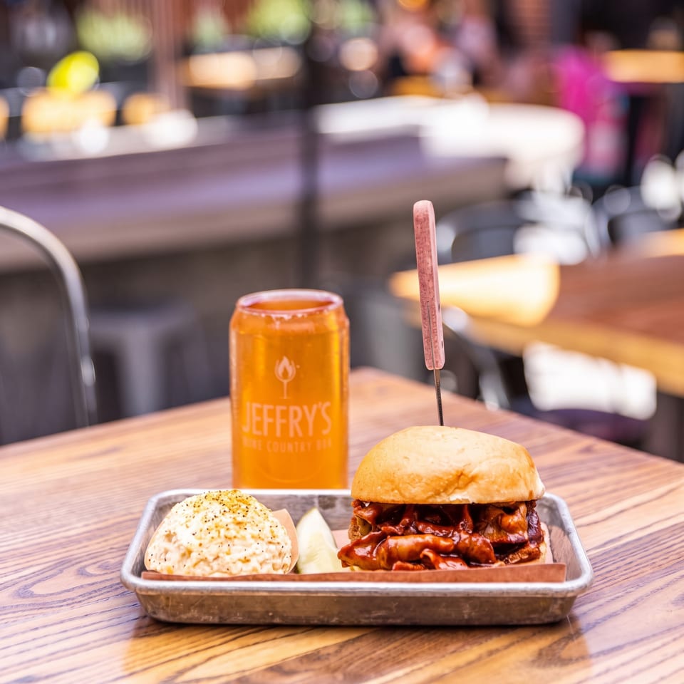 Barbecue burger with slaw and fries served on a metal tray beside a branded orange drink in a warmly lit restaurant.