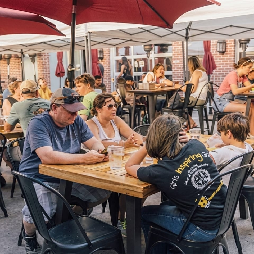 Patrons enjoy drinks and conversation at outdoor tables under red and white umbrellas on a sunny day.
