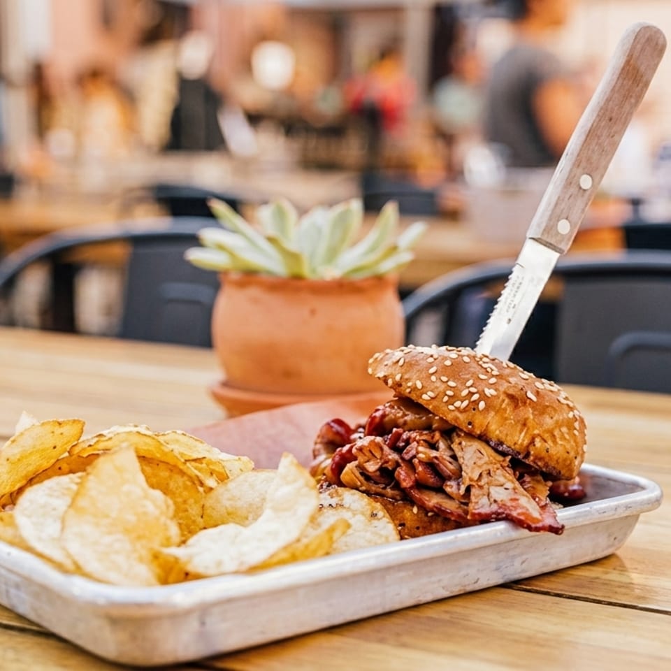 Pulled pork sandwich with chips on a metal tray at an outdoor restaurant table.