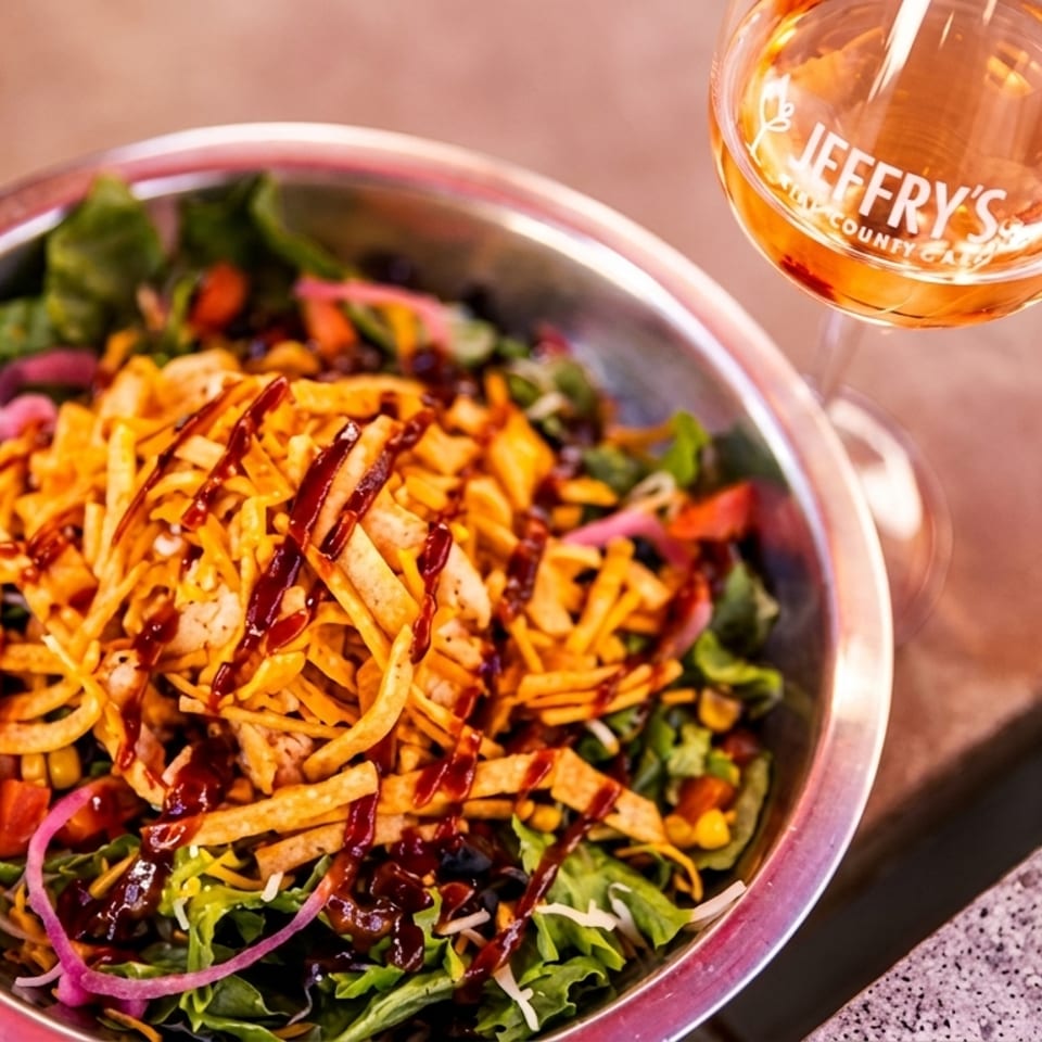 Loaded salad with greens, vegetables, crispy tortilla strips, shredded cheese, and barbecue drizzle beside a Jeffrey’s Country-style drink.