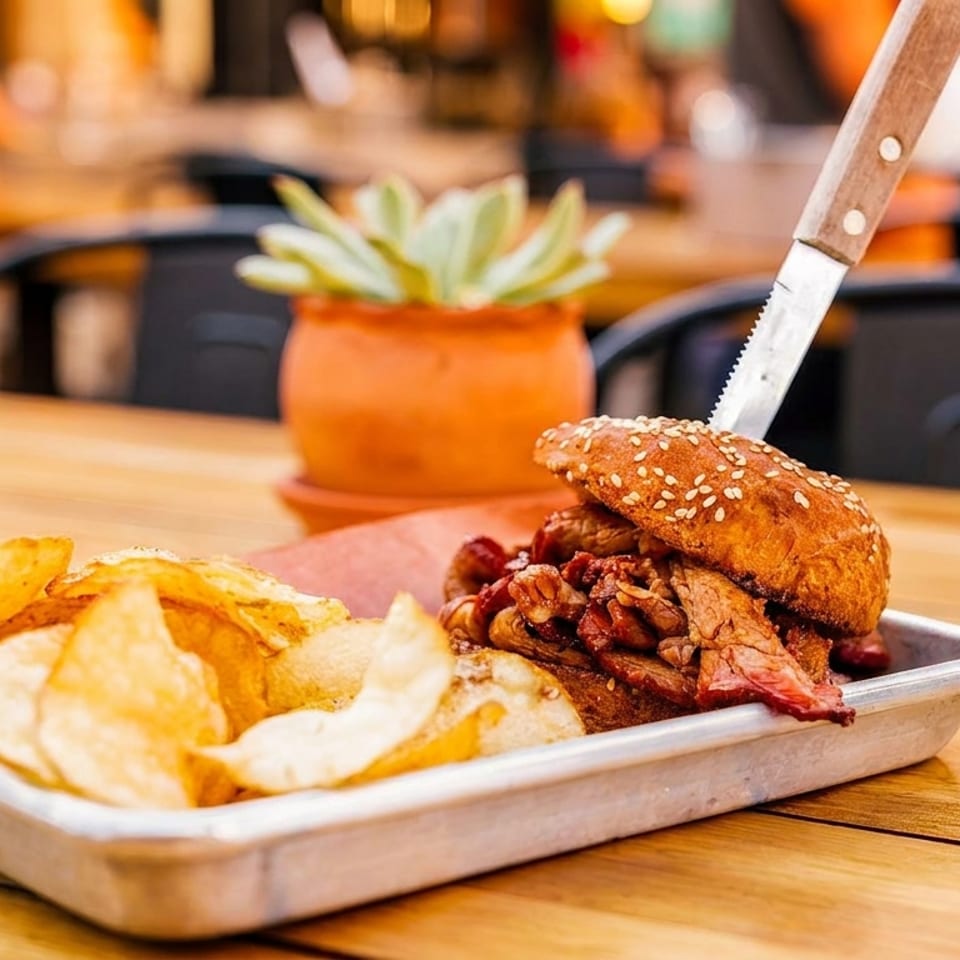 A BBQ sandwich with a knife in it, served with potato chips on a metal tray.