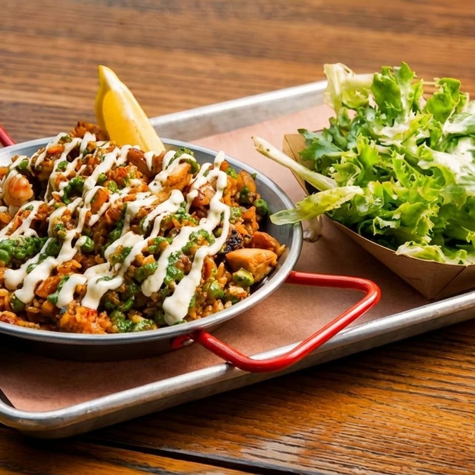 Skillet of seasoned rice, vegetables, and sauce with lemon wedge beside a fresh green salad on a metal tray.