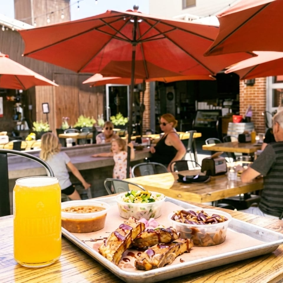 A tray of BBQ ribs, beans, and coleslaw with a beer on an outdoor patio with diners.