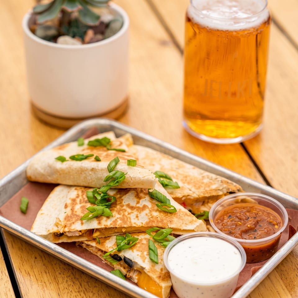 Toasted quesadillas topped with green onions, served with white and red dipping sauces, pickles, and a glass of beer.